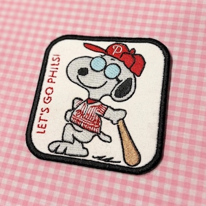 May include: Embroidered patch featuring Snoopy in a Phillies baseball uniform, holding a bat. Snoopy wears a red cap and glasses. The patch has the text "LET'S GO PHILS!" in red on a white background with a black border.