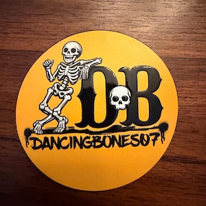 Peut inclure: Autocollant rond jaune avec un squelette dansant noir et blanc. Les lettres "DB" sont également noires, avec un petit crâne. Le texte "DANCINGBONES07" est imprimé en bas.