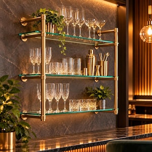 Puede incluir: Una estantería de pared de tres niveles con tubos de metal dorado cepillado y estantes de vidrio transparente. Los estantes contienen varios cristalería, incluyendo copas de vino, vasos, una coctelera dorada, un medidor y plantas pequeñas.