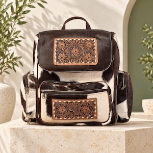 May include: A backpack crafted from black and white cowhide featuring a top handle and two side pockets with zippers. The front flap and the lower zippered pocket are each adorned with a rectangular, tan leather panel featuring intricate floral tooling and white stitched edges.