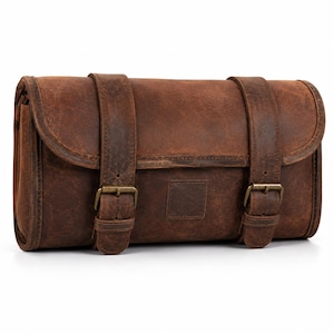 Può includere: Una borsa rettangolare in pelle marrone con finitura vissuta, dotata di una patta di chiusura fissata da due cinghie verticali in pelle con fibbie in metallo color ottone anticato. La borsa presenta una texture liscia e opaca e un design.
