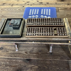 Peut inclure: Deux bouliers en bois avec des perles marron foncé, un plateau rectangulaire noir contenant une petite pierre à encre et un bâton d'encre, et un coussin rectangulaire bleu avec un motif floral blanc, disposés sur une surface en bois vieilli.