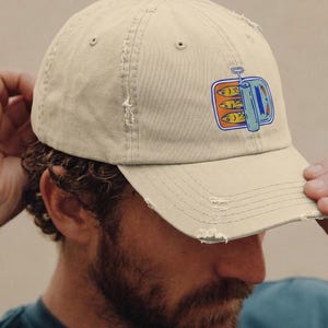 May include: Tan baseball cap with a distressed brim. The cap features a colorful graphic of sardines in a can. The design includes orange, blue, and yellow accents. The cap is worn by a person.