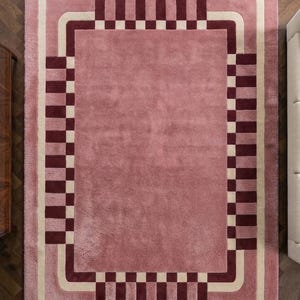 May include: A rectangular area rug featuring a solid dusty rose center surrounded by a cream-colored border and a dark burgundy checkered pattern. The rug has a plush, textured surface with rounded corners on the inner border design.