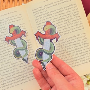 May include: Two die-cut stickers featuring a silver dagger with a red hilt wrapped in a green snake. The stickers are held against an open book page titled In the Witch's House, surrounded by pink and white flowers on a dark pink surface.
