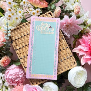 May include: A rectangular notepad with a light blue grid background and a pink border featuring floral patterns. The top section displays the text "Side Quests Unlocked" in a decorative font, followed by a vertical list of checkboxes for tasks.