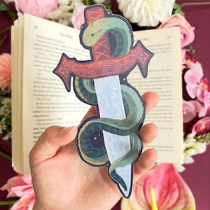 May include: A die-cut sticker featuring a silver sword with a light blue lightning pattern on the blade, wrapped in a green snake. A dark red, ornate banner crosses the sword hilt. The sticker has a dark blue outline and a matte finish.