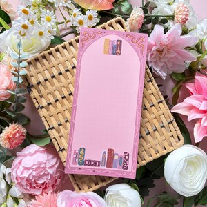 May include: A rectangular pink notepad featuring a grid pattern, an arched border with snake illustrations, and a decorative base of colorful book spines. The notepad is positioned on a woven tan tray surrounded by various artificial flowers.