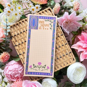 May include: A rectangular notepad with a grid pattern, featuring a blue and purple border with floral accents and the text "Once upon a..." in a decorative font. The design includes a large stylized letter O and pink flower illustrations.
