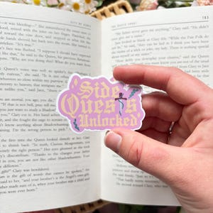 May include: A die-cut sticker with a light purple background and a wavy white border. The text reads Side Quests Unlocked in a stylised gold font. A small dark blue sword icon is positioned vertically between the words Quests and Unlocked.