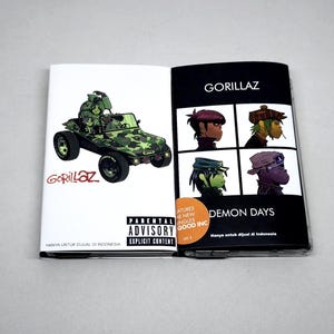 May include: An open Gorillaz Demon Days CD case featuring a green camouflage buggy on the left panel and four illustrated character portraits on the right panel. The cover includes the text Gorillaz, Demon Days, and a Parental Advisory label.