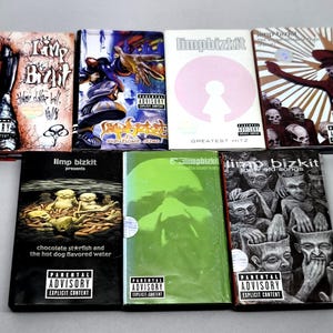 Pode incluir: Uma coleção de CDs de música do Limp Bizkit com várias capas de álbuns. As capas apresentam diversas obras de arte, incluindo ilustrações, logotipos da banda e o selo Parental Advisory. Os CDs estão dispostos sobre uma superfície cinza claro.