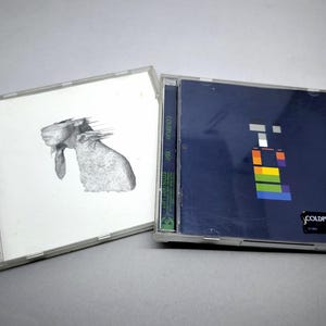 May include: Two plastic CD jewel cases. The left case features a white cover with a black and white sketch of a goat. The right case has a dark blue cover with a colourful block graphic and the text COLDPLAY X&Y printed in the bottom corner.
