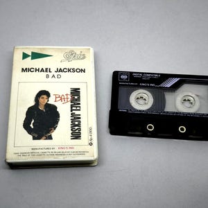 Puede incluir: Una cinta de cassette vintage y su estuche del álbum "Bad" de Michael Jackson. El estuche es blanco con el título del álbum y el nombre del artista. El cassette es negro con bobinas de cinta visibles.