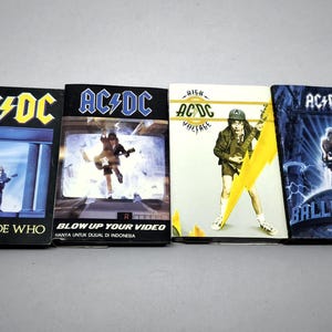May include: Four AC/DC music cassette tapes featuring album covers for Who Made Who, Blow Up Your Video, High Voltage, and Ballbreaker. Each rectangular case displays the band logo and specific artwork, including illustrations of a guitarist.