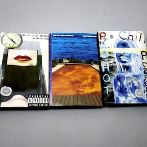 Pode incluir: São exibidos três álbuns de CD do Red Hot Chili Peppers. As capas dos álbuns apresentam diversas obras de arte, incluindo um relógio, lábios e imagens abstratas. Os álbuns são intitulados "Greatest Hits" e "Californication". Um aviso para os pais é visível.