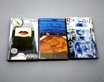 May include: Three Red Hot Chili Peppers CD albums are shown. The album covers display varied artwork, including a clock, lips, and abstract designs. The albums are titled "Greatest Hits" and "Californication". A parental advisory warning is visible.