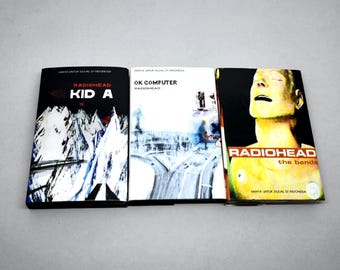 May include: Three Radiohead music albums arranged side by side. The first is Kid A with a black and white mountain graphic. The second is OK Computer with a white cover and abstract imagery. The third is The Bends featuring a yellow face graphic.