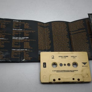 May include: A beige cassette tape and its unfolded inlay card. The tape is labelled "Guns N' Roses". The inlay is black with song lyrics in a brown font. A retro music item, reminiscent of the 1980s and 1990s.