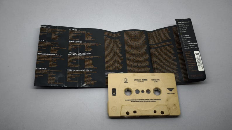 May include: A beige cassette tape and its unfolded inlay card. The tape is labelled "Guns N' Roses". The inlay is black with song lyrics in a brown font. A retro music item, reminiscent of the 1980s and 1990s.