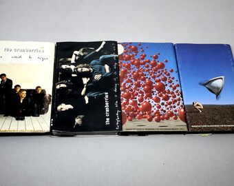 May include: Four music cassette tapes featuring The Cranberries albums. The covers display band photography, red spheres, and a desert landscape with a floating object. Text includes "the cranberries", "no need to argue", and "everybody else is doing it".