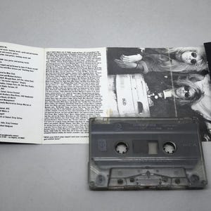 May include: A retro audio cassette tape with its accompanying artwork and liner notes. The cassette is transparent, revealing its internal mechanisms. The cover art displays a black and white photograph of a person wearing sunglasses. The liner notes are printed on white paper.