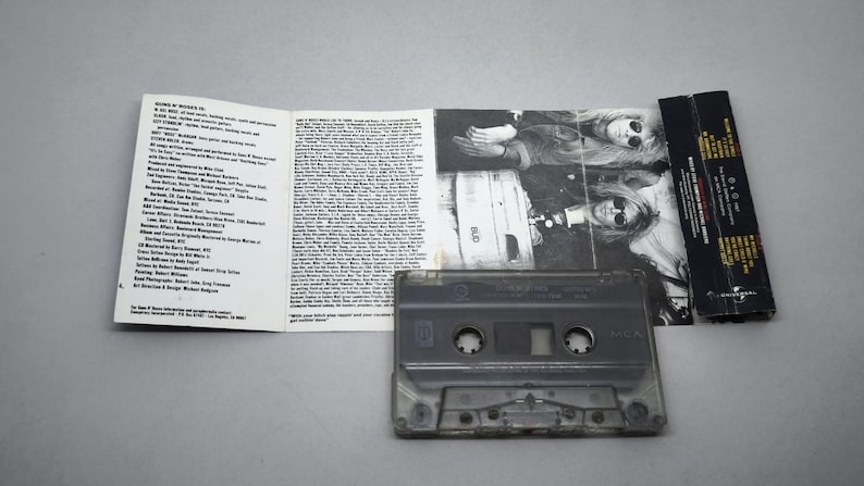 May include: A retro audio cassette tape with its accompanying artwork and liner notes. The cassette is transparent, revealing its internal mechanisms. The cover art displays a black and white photograph of a person wearing sunglasses. The liner notes are printed on white paper.