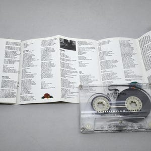 May include: A transparent cassette tape with black internal components and white spools, alongside an open booklet filled with song lyrics. The booklet features a yellow and red border and black text on a white background.