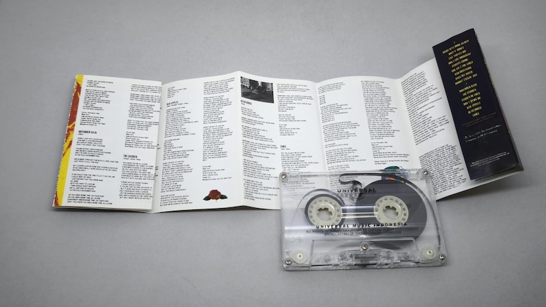 May include: A transparent cassette tape with black internal components and white spools, alongside an open booklet filled with song lyrics. The booklet features a yellow and red border and black text on a white background.