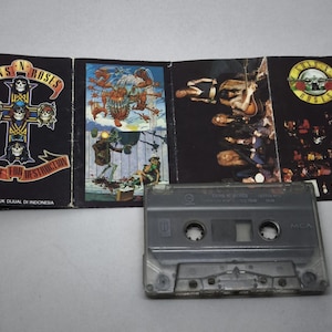 May include: A Guns N' Roses cassette tape and its cover art. The cover art features the band's logo and album art. The cassette is grey and has a clear plastic window. The album cover is black with colourful illustrations.