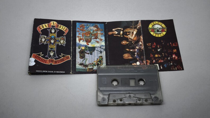 May include: A Guns N' Roses cassette tape and its cover art. The cover art features the band's logo and album art. The cassette is grey and has a clear plastic window. The album cover is black with colourful illustrations.