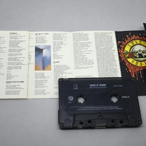 May include: A black cassette tape and the accompanying booklet for Guns N' Roses' album, "G N' R Lies." The album art features a skull with roses and the band's name. The booklet is open, displaying song lyrics.