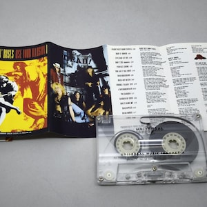 May include: A Guns N' Roses "Use Your Illusion I" cassette tape with its booklet. The cassette is transparent, showing the tape inside. The booklet has album art and song lyrics. The cover art features a figure in front of a yellow and orange background.