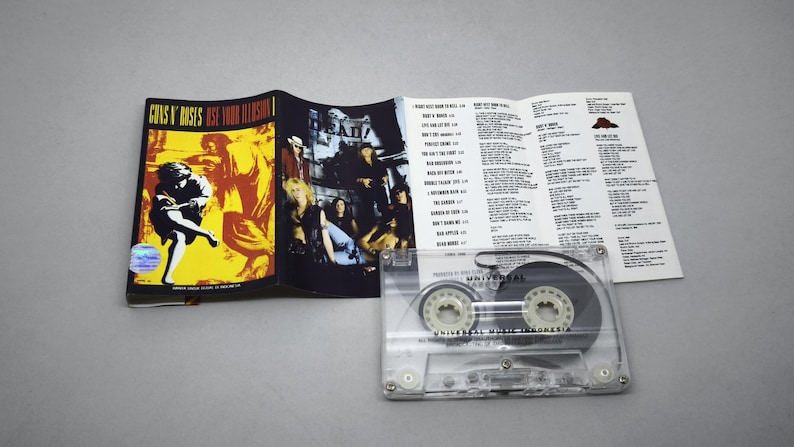 May include: A Guns N' Roses "Use Your Illusion I" cassette tape with its booklet. The cassette is transparent, showing the tape inside. The booklet has album art and song lyrics. The cover art features a figure in front of a yellow and orange background.