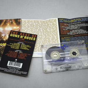 May include: A clear cassette tape with a black and white label, and a folded booklet with the text "THE BEST OF GUNS N' ROSES LIVE" and a concert photo. The cassette case is transparent.