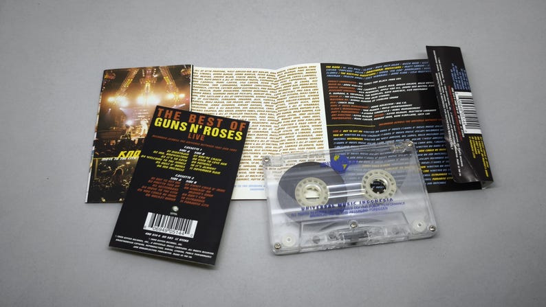 May include: A clear cassette tape with a black and white label, and a folded booklet with the text "THE BEST OF GUNS N' ROSES LIVE" and a concert photo. The cassette case is transparent.
