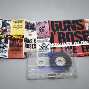 May include: A Guns N' Roses cassette tape with a folded promotional poster. The poster features concert dates, skull graphics, and the band's name in red and black. The cassette is clear with a black tape.