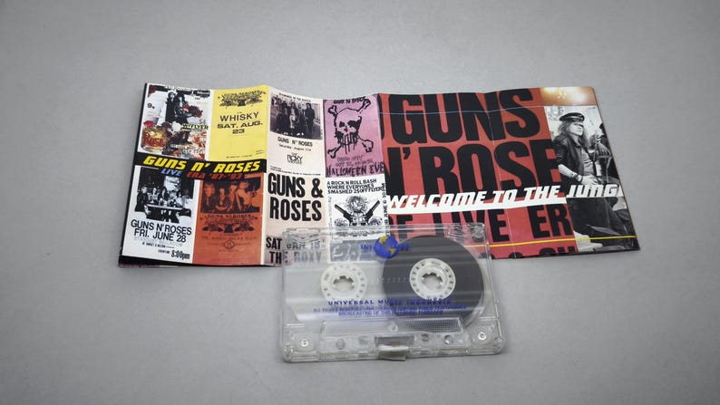 May include: A Guns N' Roses cassette tape with a folded promotional poster. The poster features concert dates, skull graphics, and the band's name in red and black. The cassette is clear with a black tape.