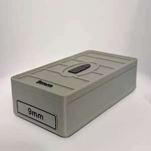 May include: A rectangular light grey storage box with a lid featuring a raised black bullet icon and the text 9mm. The front side of the box displays a rectangular label with the text 9mm in black lettering. The surface has a visible layered texture.
