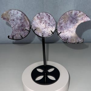 May include: A triple moon display featuring three polished lepidolite stone carvings in crescent and circular shapes. The stones are mounted on a black metal stand with a circular base, showcasing purple and white mottled mineral patterns.