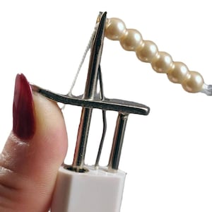 May include: A metal sewing tool with a white base and a vertical needle mechanism. A thin white thread passes through the needle eye, which is currently threaded with a string of six small, round, cream-colored faux pearls in a linear arrangement.