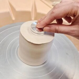 Puede incluir: Una pieza cilíndrica de arcilla de color beige claro centrada en un torno de alfarero circular de metal. Una pequeña herramienta de metal redonda de color plateado se presiona firmemente sobre la superficie superior plana de la arcilla.