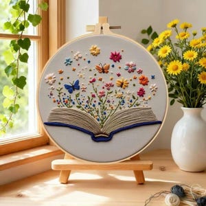 Può includere: Un telaio da ricamo rotondo con un libro aperto ricamato con fiori e farfalle colorate. Il design utilizza colori come blu, rosa, arancione e giallo su uno sfondo grigio chiaro. Il telaio è esposto su un piccolo cavalletto di legno.