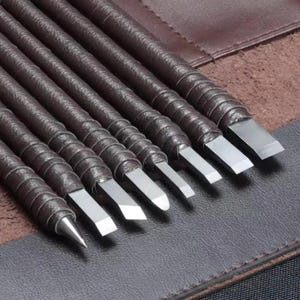 May include: A set of eight leather-wrapped carving tools with stainless steel blades. The tools are arranged in a brown leather case, with the blades facing up. The handles are dark brown and spiraled, and the blades are various shapes.