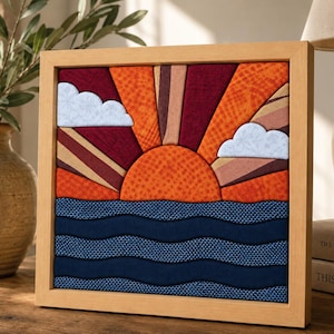 May include: A square framed fabric wall art piece featuring a sunrise over the ocean. The scene includes a textured orange sun, radiating beams of maroon and orange fabric, and two light blue clouds. The sea is depicted with wavy dark blue fabric.