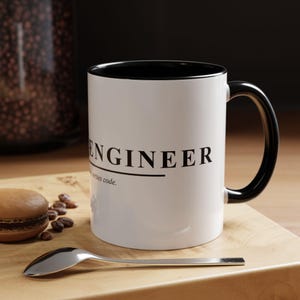 May include: White ceramic mug with a black interior and handle. The mug features the word "ENGINEER" in black, with the phrase "...writes code." below. A spoon, a macaron, and coffee beans are on a wooden surface.