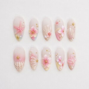 Könnte beinhalten: Ein Set aus zehn künstlichen Nägeln, jedes mit einem einzigartigen Design. Sie zeigen rosa und weiße Farbverläufe, Muscheln, Seesterne und florale Akzente. Goldfarbene Verzierungen verleihen der Nagelkunst einen Hauch von Glanz.