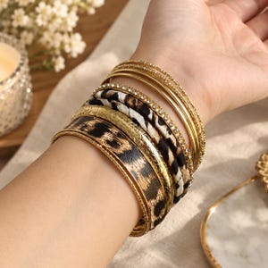 May include: A stack of multiple bangles including thin gold-tone metal bands, a band encrusted with small clear crystals, a band with a textured gold finish, and two wider bands featuring a brown and black leopard print pattern on a smooth surface.