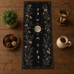 May include: A rectangular black table runner featuring a central vertical column of moon phases and a full moon, flanked by detailed botanical illustrations of wildflowers, daisies, and lavender in muted tones of pink, white, and green.