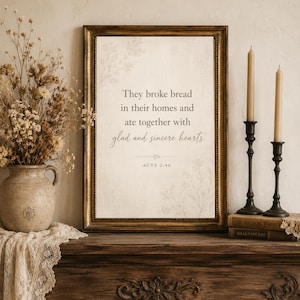 May include: A framed print with the text "They broke bread in their homes and ate together with glad and sincere hearts. ACTS 2:46." The frame is gold, and the print is displayed on a wooden surface with candles and books.